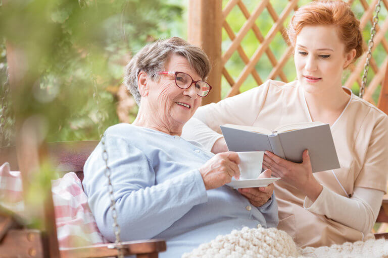 happy elderly woman drinking tea while nurse reading book in the garden