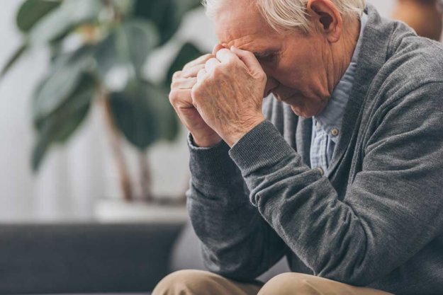 selective focus of upset retired man holding head while sitting on sofa