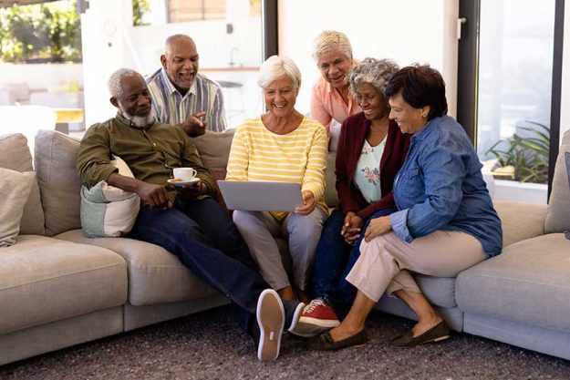 biracial senior woman using laptop with multiracial friends while sitting on sofa in nursing home