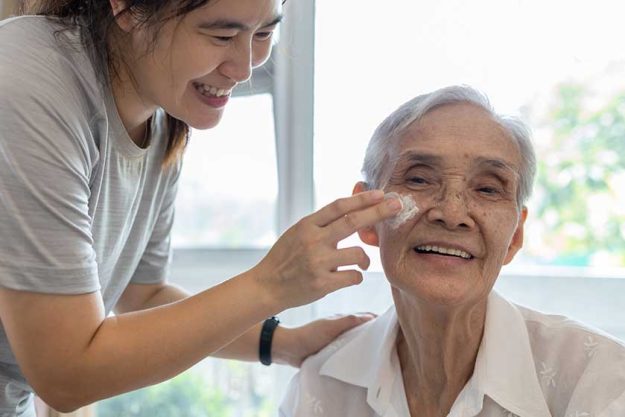 female caregiver assisting to apply sunscreen lotion on the face of senior woman