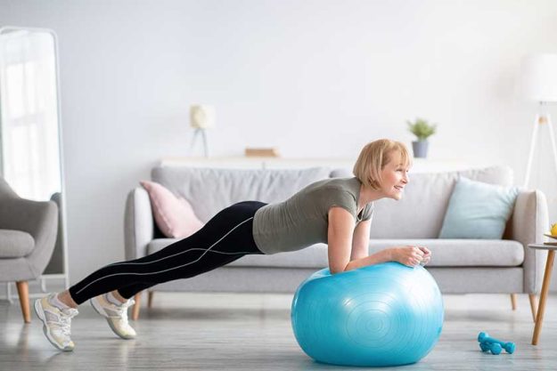 athletic mature woman standing in plank on fitness ball working out core muscles at home