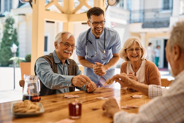 happy senior friends and male nurse laughing while playing cards