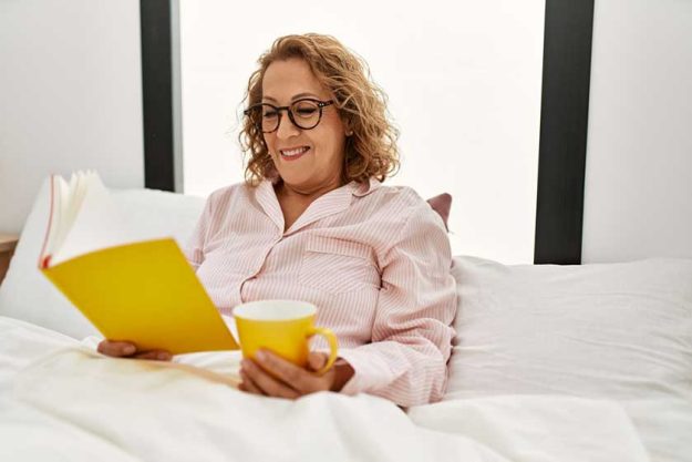 happy senior woman reading book and drinking coffee in bed at home