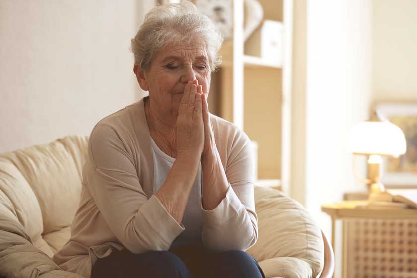 praying elderly woman sitting on lounge at home
