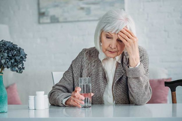 portrait of grey hair woman with headache sitting at table with pills and glass of water at home
