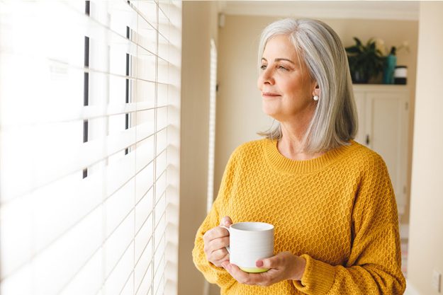 thoughtful caucasian senior woman standing looking out window home coffee