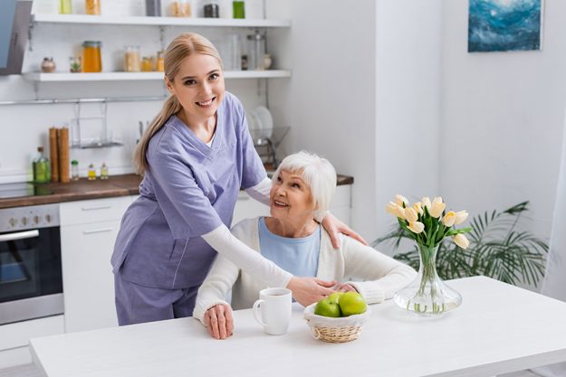 young nurse smiling camera while embracing happy aged woman kitchen
