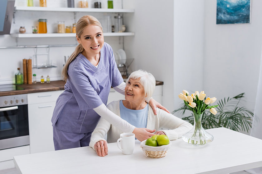 young nurse smiling camera while embracing happy aged woman kitchen