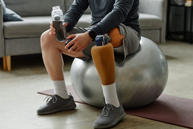 close unrecognizable man prosthetic leg sitting fitness ball holding water