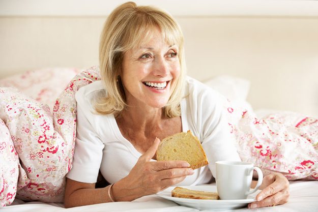 senior woman snuggled under duvet eating breakfast