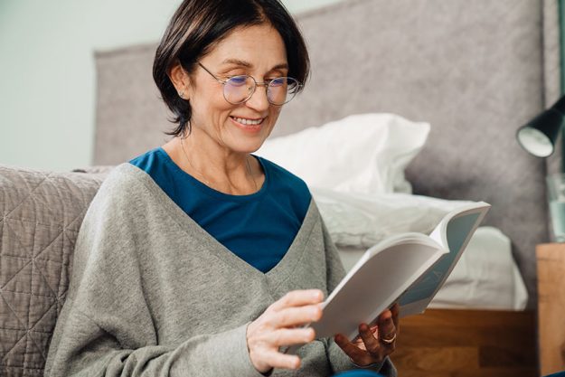 white mature woman eyeglasses reading book while sitting floor home