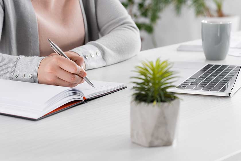 cropped view freelancer writing notebook laptop plant blurred foreground cropped view freelancer writing notebook laptop plant blurred foreground