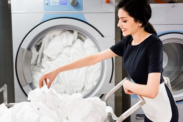 cheerful maid looking bed sheets while standing washing machines