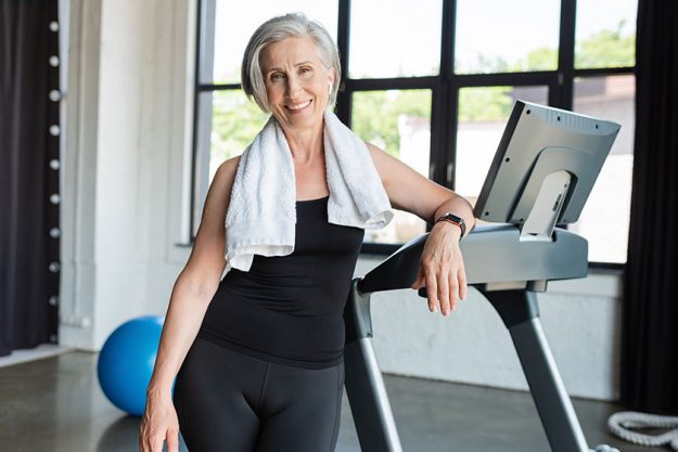 Happy senior woman white towel shoulders standing next treadmill