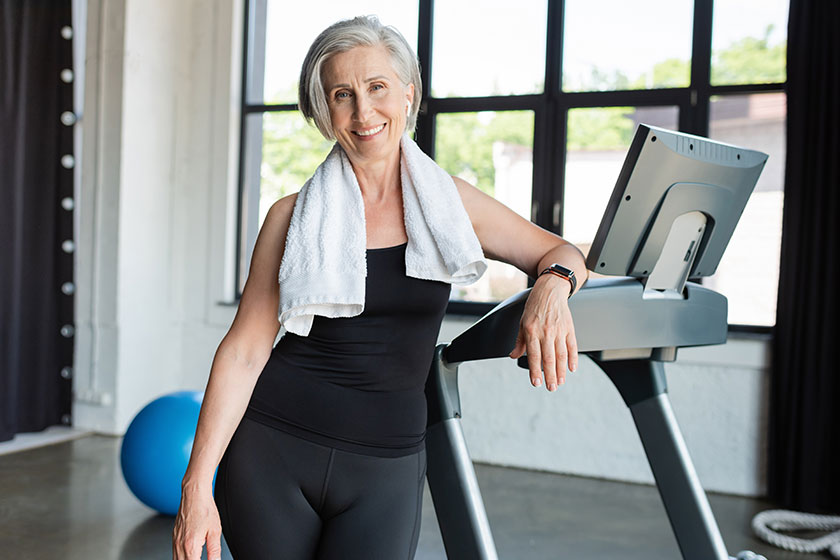 Happy senior woman white towel shoulders standing next treadmill