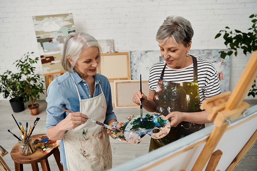 Two women stand together art studio holding paintbrushes palette engaged