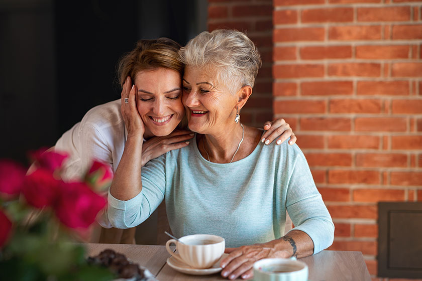Happy senior mother embracing with adult daughter when sitting and having coffee indoors at home