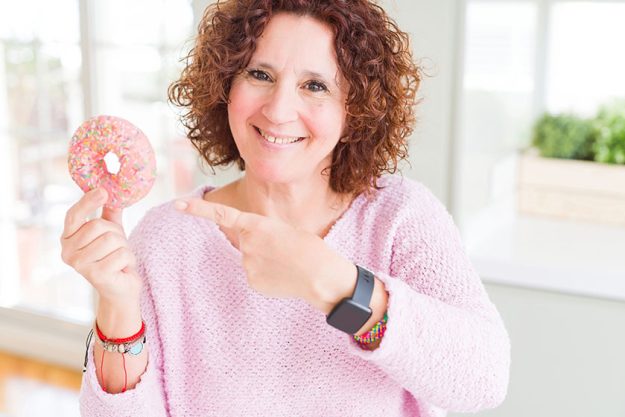Senior woman eating pink sugar donut very happy pointing hand