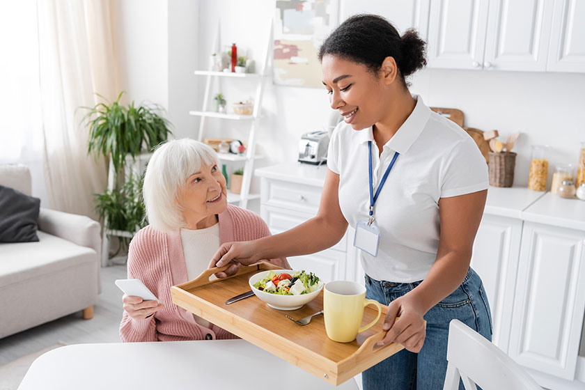 Happy multiracial social worker holding tray lunch senior woman grey Happy multiracial social worker holding tray lunch senior woman grey