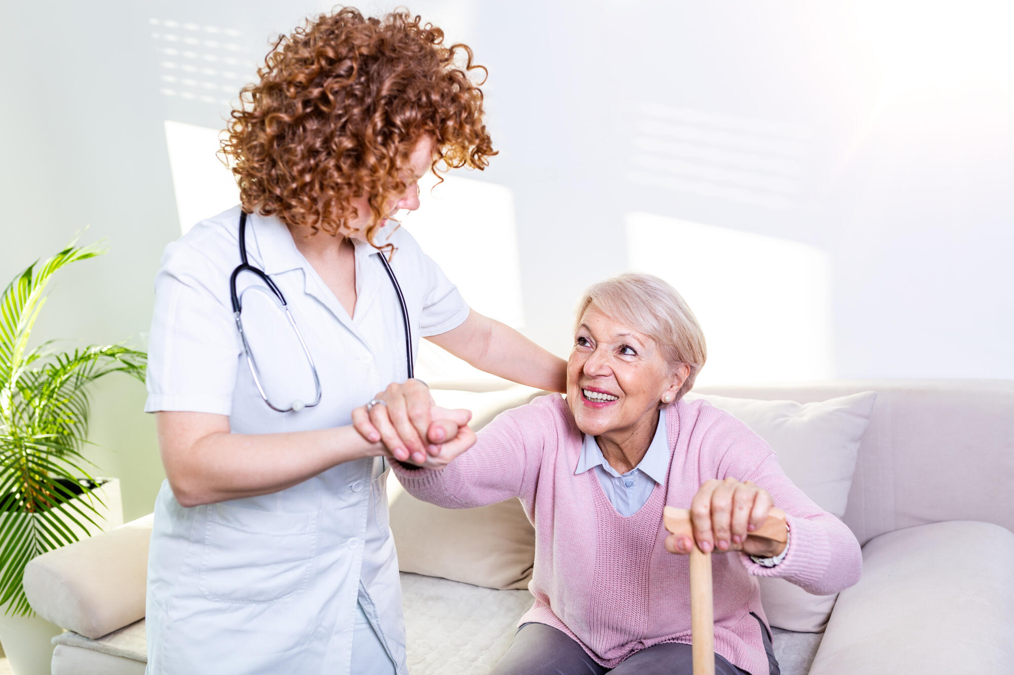 Female caregiver helping senior woman get up from couch in living room. Smiling nurse assisting senior woman to get up. Caring nurse supporting patient while getting up from sofa