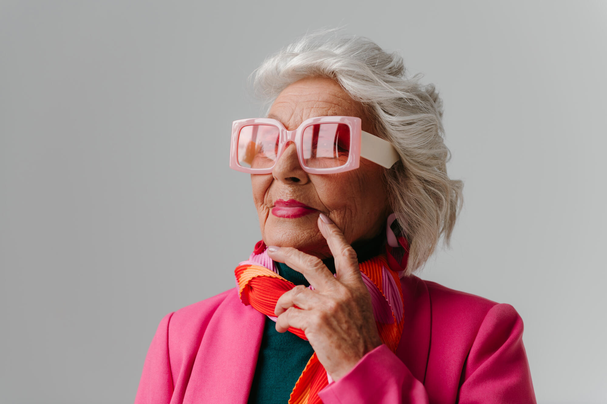 Elegant elderly woman wearing fashionable retro styled eyeglasses and radiating self love on grey background