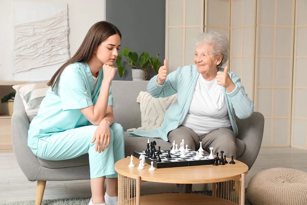 Young caregiver senior woman playing chess home