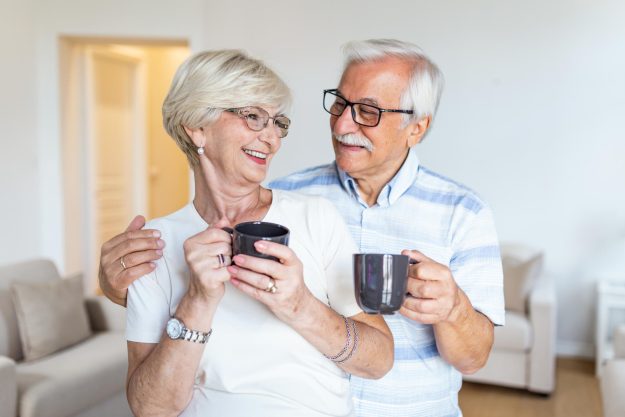 Happy senior couple standing in the living room. Looking each other smilling and holding cup of coffee or tea. Elderly husband hugging his lovely wife