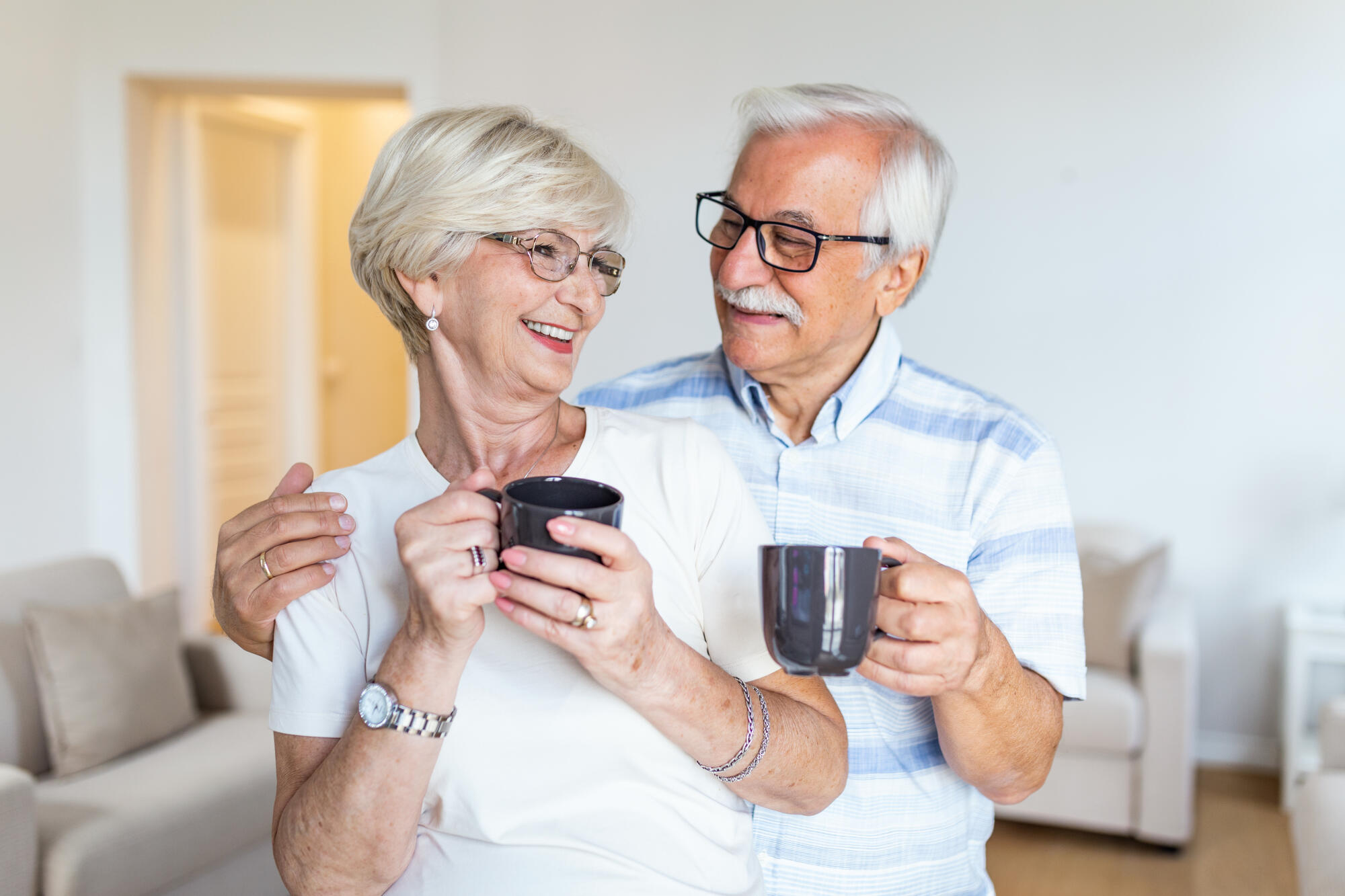 Happy senior couple standing in the living room. Looking each other smilling and holding cup of coffee or tea. Elderly husband hugging his lovely wife Happy senior couple standing in the living room. Looking each other smilling and holding cup of coffee or tea. Elderly husband hugging his lovely wife