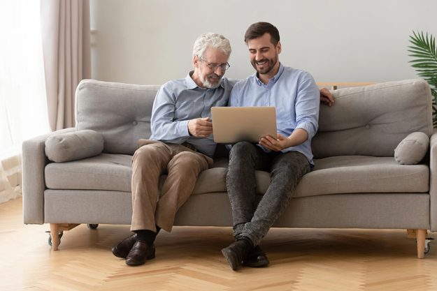 Father and grown up son sit together on sofa with computer Father and grown up son sit together on sofa with computer