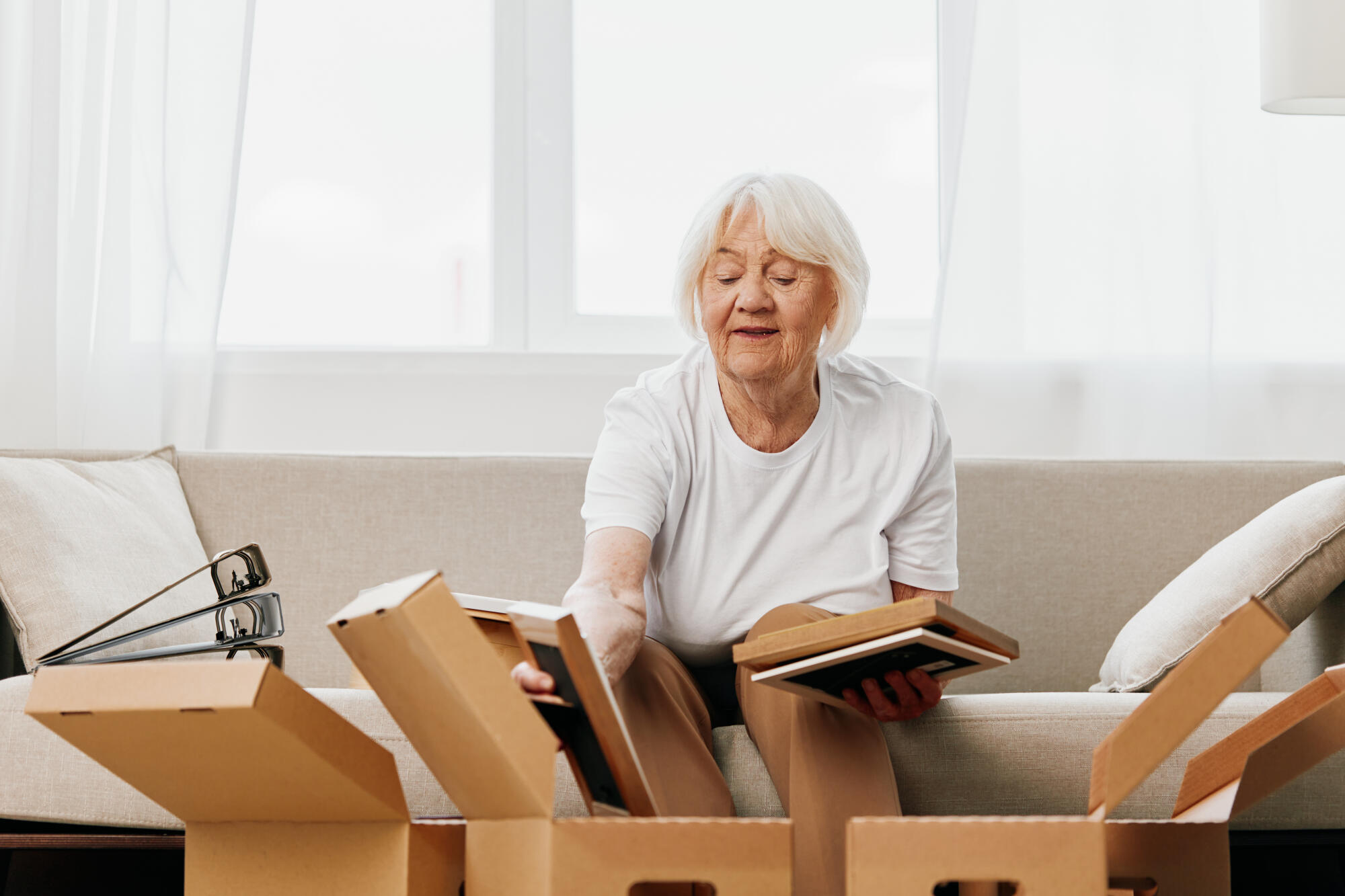 elderly woman sits on a sofa at home with boxes. collecting things with memories albums with photos and photo frames moving to a new place cleaning things and a happy smile. Lifestyle retirement. elderly woman sits on a sofa at home with boxes. collecting things with memories albums with photos and photo frames moving to a new place cleaning things and a happy smile. Lifestyle retirement.