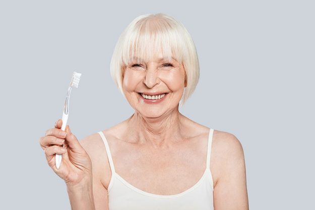 Smiling senior woman holding toothbrush standing grey background looking camera Smiling senior woman holding toothbrush standing grey background looking camera