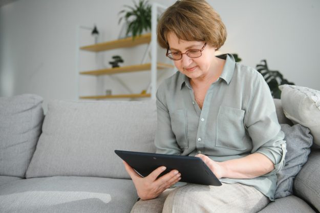 Retirement Leisure. Smiling Elderly Woman Resting With Digital Tablet On Couch At Home, Browsing Internet Or Watching Movies Retirement Leisure. Smiling Elderly Woman Resting With Digital Tablet On Couch At Home, Browsing Internet Or Watching Movies