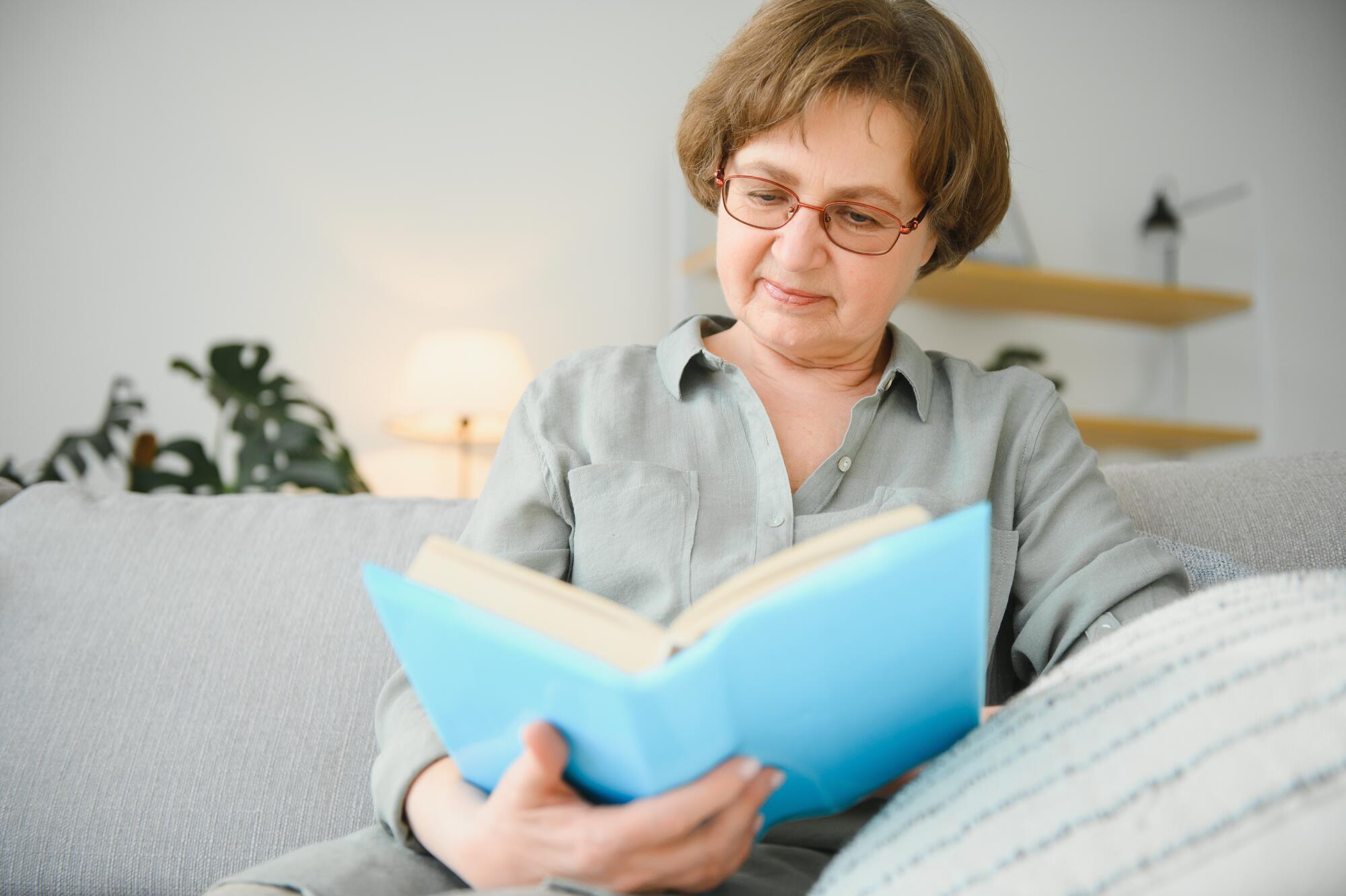 Restful reading. Portrait of thoughtful aged woman reading favorite literature at cozy home. She is lying on pillows on comfortable sofa. Legs are covered with soft blanket.