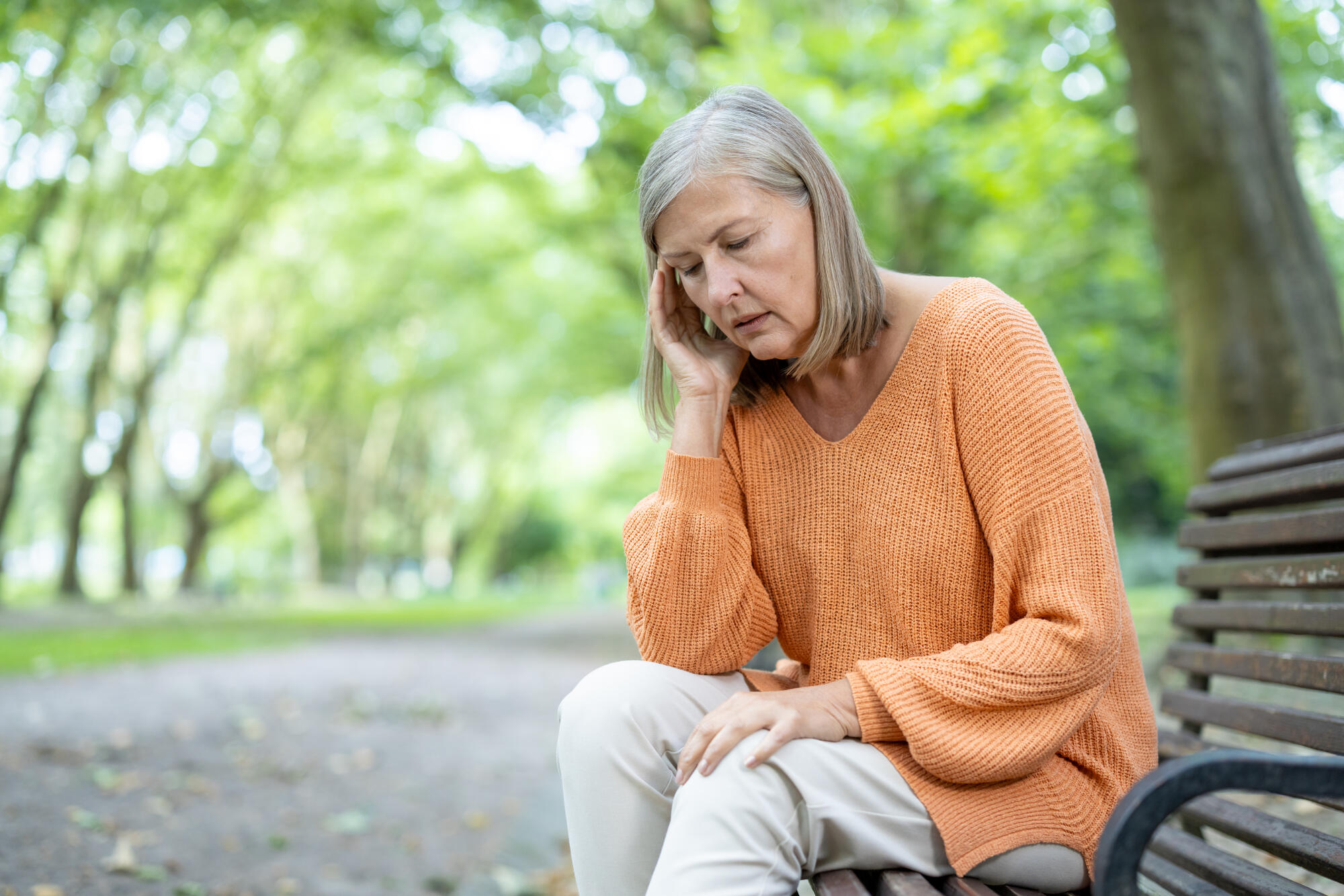 Thoughtful elderly woman sitting on park bench wearing orange sweater, enjoying peaceful outdoor environment, representing reflection and solitude in a tranquil green setting.