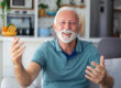 Smiling senior man wave to camera having video call on laptop, happy elderly male sit on couch at home talk using modern technologies and wireless connection