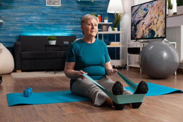 Retirement senior woman sitting on yoga mat stretching legs muscles using stretch elastic band Retirement senior woman sitting on yoga mat stretching legs muscles using stretch elastic band