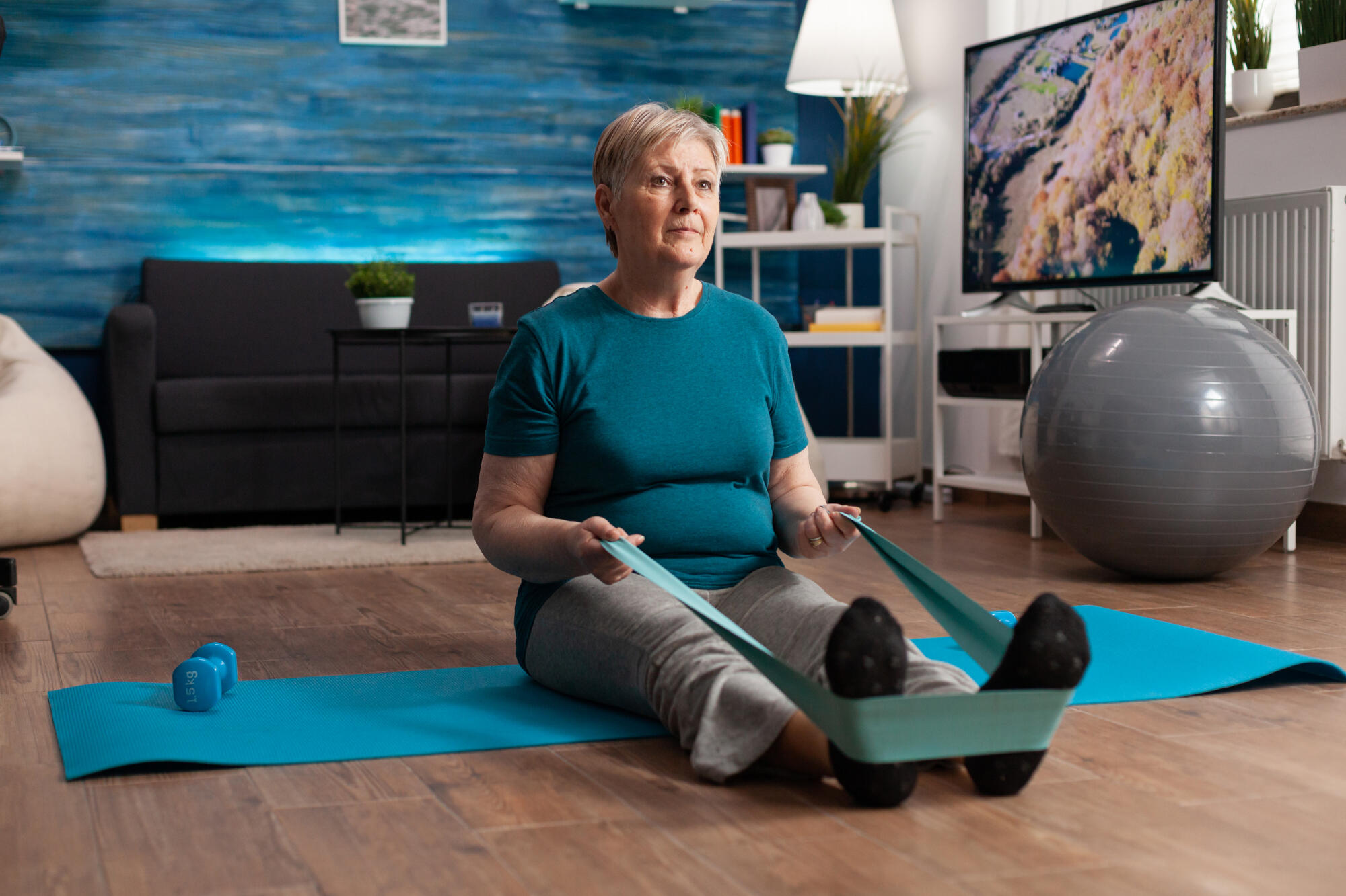 Retirement senior woman sitting on yoga mat stretching legs muscles using stretch elastic band