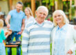 Enjoying quality time with family. Happy senior couple bonding to each other and looking at camera while other members of family barbecuing food in the background
