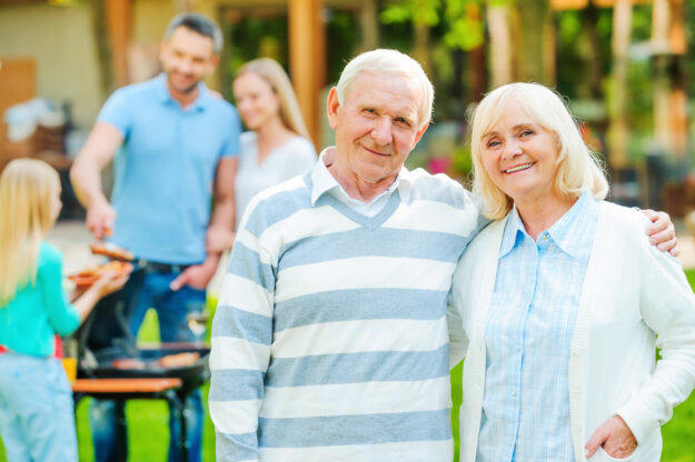 Enjoying quality time with family. Happy senior couple bonding to each other and looking at camera while other members of family barbecuing food in the background