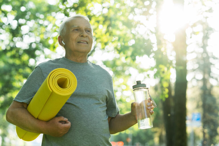 Active elderly man with a exercise mat and bottle of water in green city park