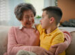 Caucasian happy family two generations elderly retired woman grandmother talking hugging boy child grandson speaking enjoying communication at home on couch grandma talk hug embrace cuddle grandchild