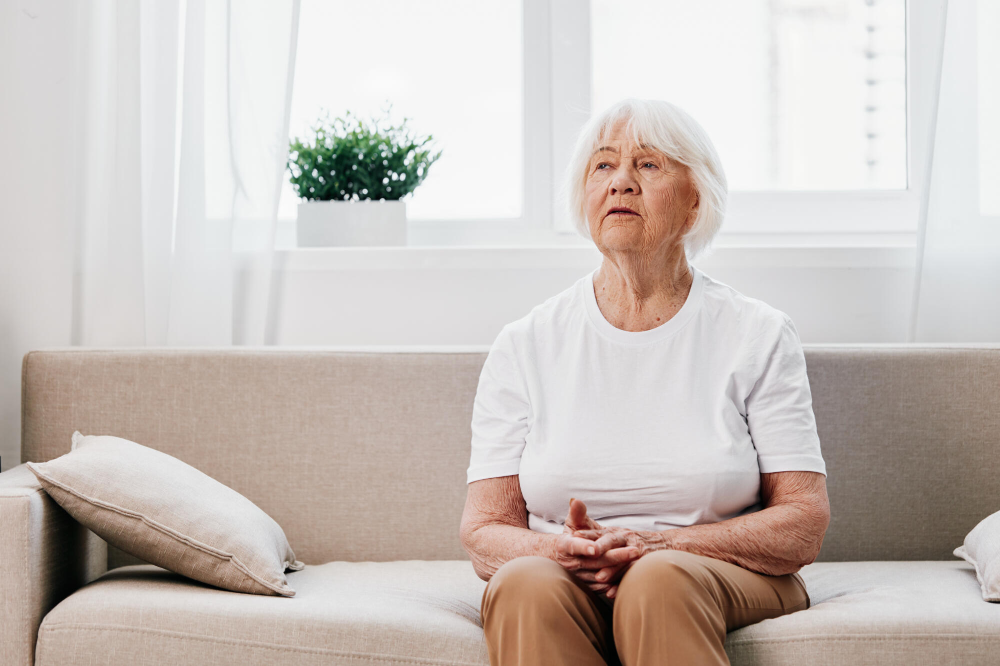 Elderly woman sits on sofa at home, bright spacious interior in old age smile, lifestyle. Grandmother with gray hair in a white T shirt and beige trousers. Elderly woman sits on sofa at home, bright spacious interior in old age smile, lifestyle. Grandmother with gray hair in a white T shirt and beige trousers.