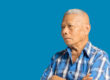 An elderly Asian man in a blue shirt arms crossed and looking aw