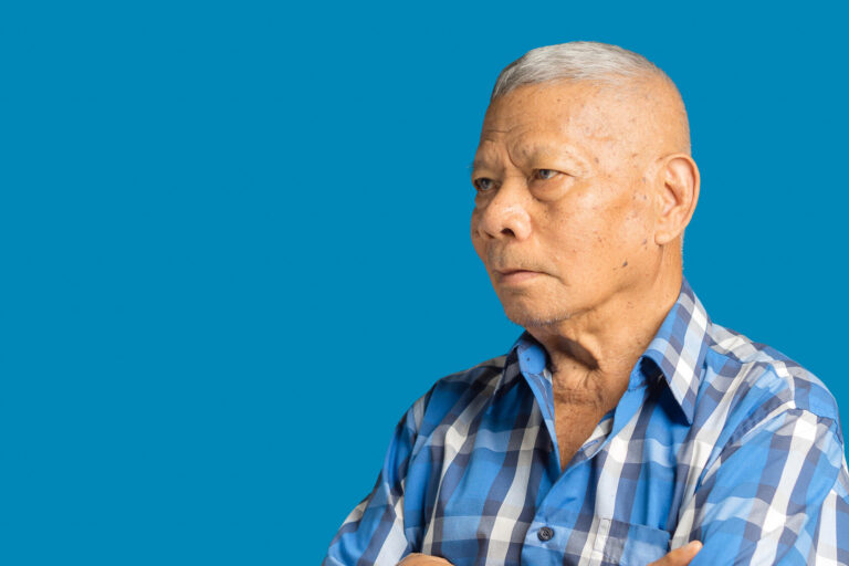 An elderly Asian man in a blue shirt arms crossed and looking aw