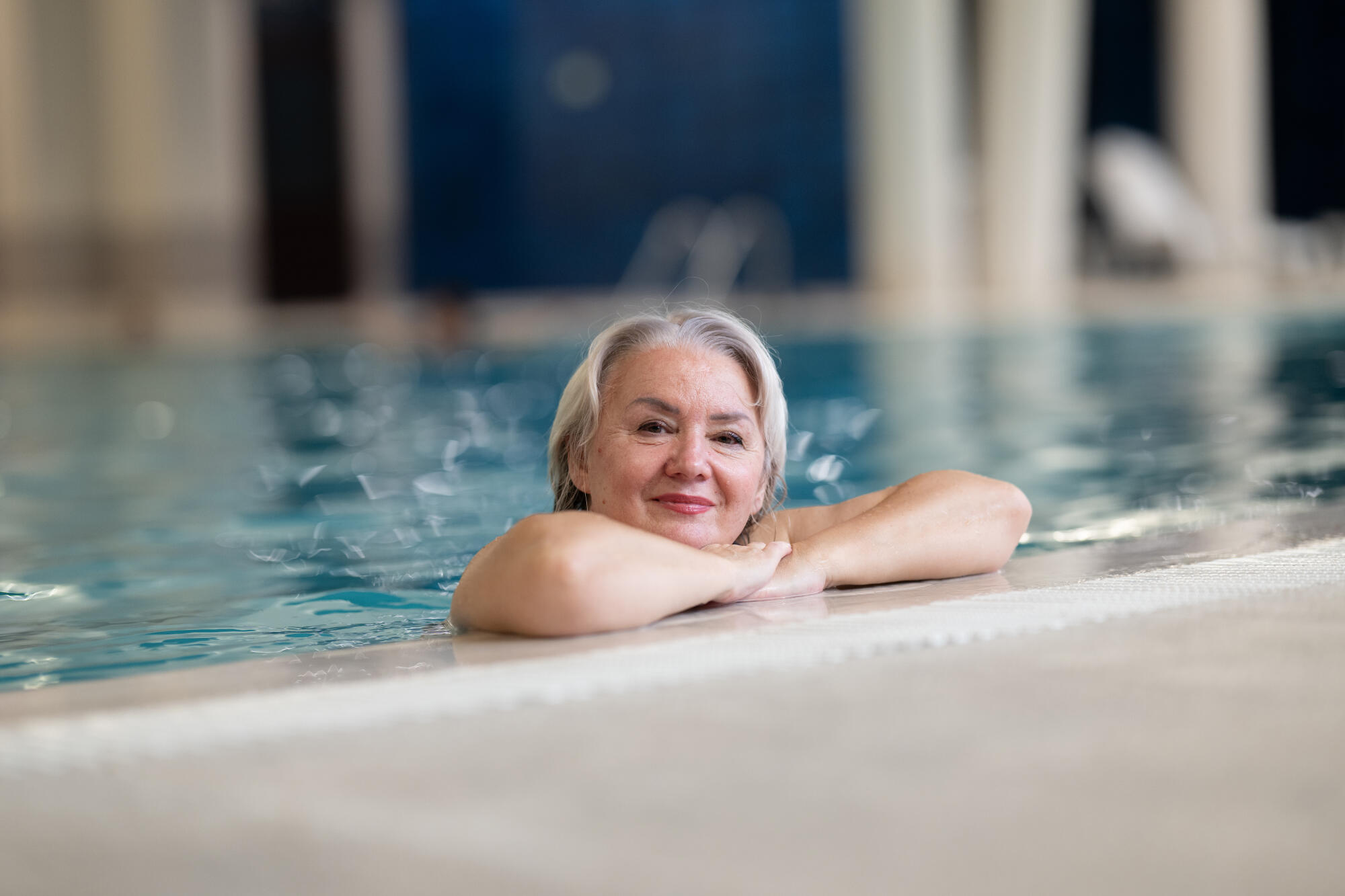 Smiling senior woman enjoying relaxation in an indoor swimming pool, leaning on the poolside. Concept of active lifestyle, wellness, health, hydrotherapy, and leisure for older adults.