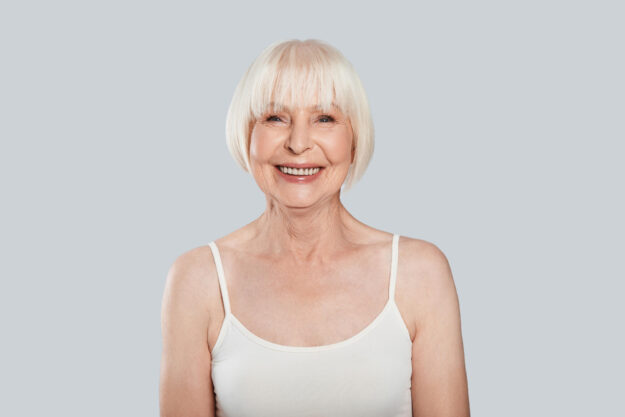 Natural beauty. Beautiful senior woman looking at camera and smiling while standing against grey background Natural beauty. Beautiful senior woman looking at camera and smiling while standing against grey background
