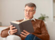 Retired man reading book sitting on couch at home