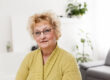 Smiling middle aged mature grey haired woman looking at camera, happy old lady in glasses posing at home indoor, positive single senior retired female sitting on sofa in living room headshot portrait