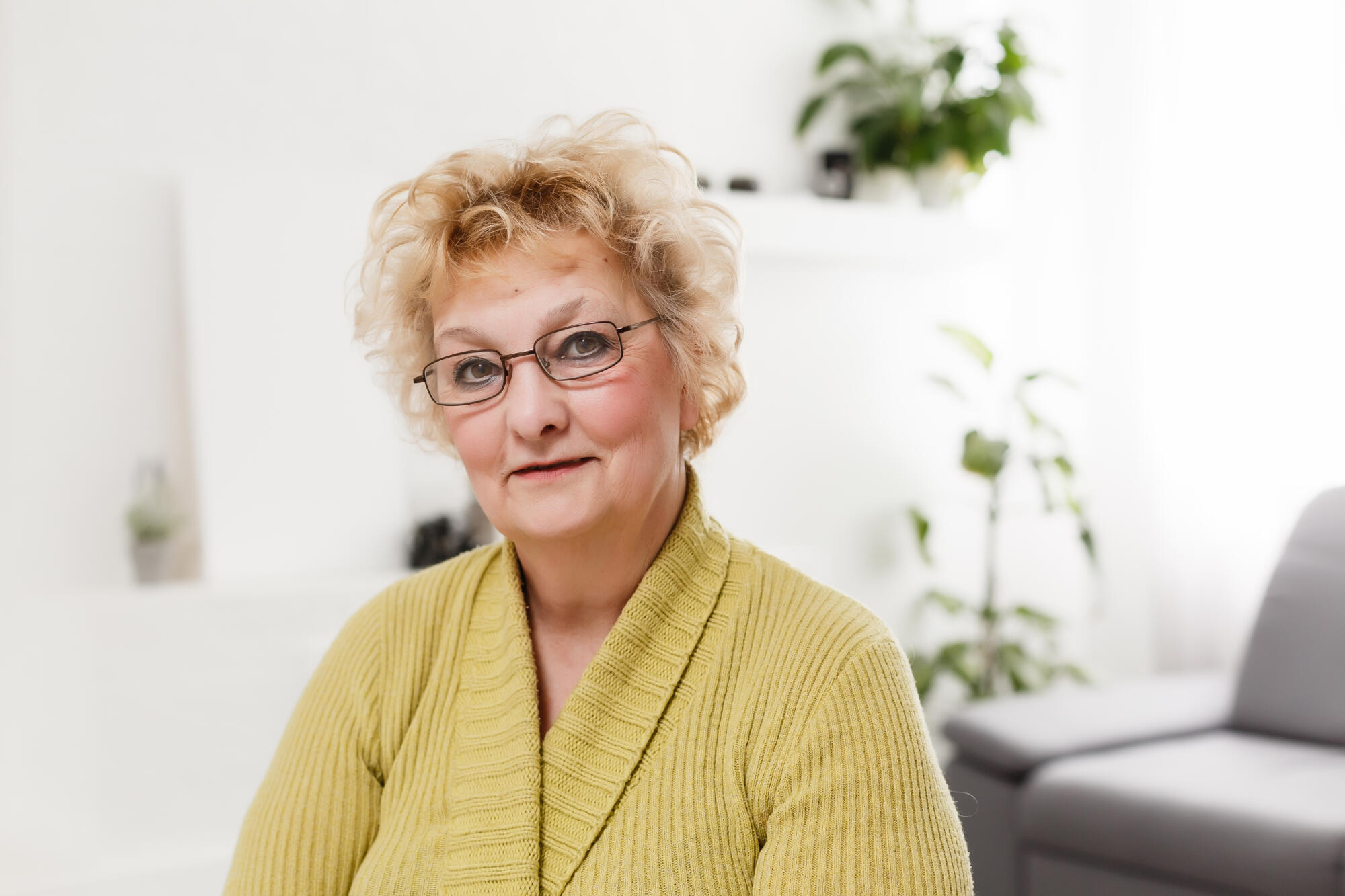 Smiling middle aged mature grey haired woman looking at camera, happy old lady in glasses posing at home indoor, positive single senior retired female sitting on sofa in living room headshot portrait