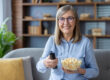 Smiling woman holding a bowl of popcorn and remote control while sitting on a cozy sofa. Enjoying leisure time at home with entertainment. Relaxed elderly lady ready to watch TV.